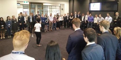 A group of people standing around in a square formation within an office building. A speaker is in the middle of the square.